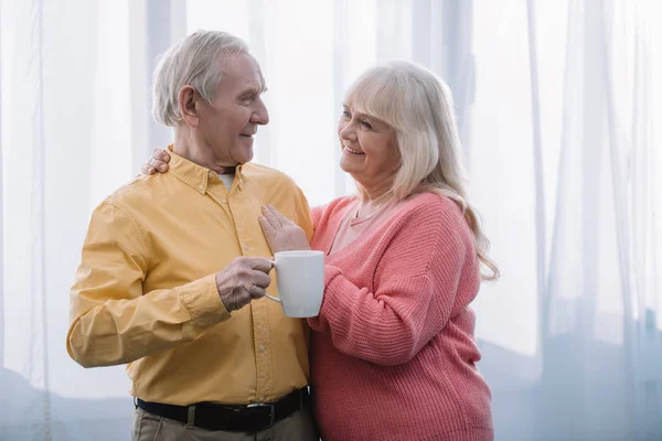 Усміхнена старша жінка з сірим волоссям дивиться на камеру і приймає чоловіка з чашкою чаю вдома — стокове фото