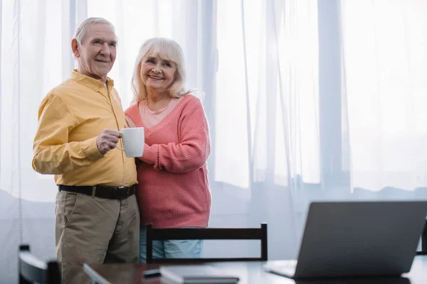 Selective focus of smiling senior couple in casual clothes holding cup of coffee at home with copy space — Stock Photo