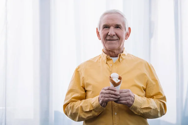 Старший мужчина смотрит в камеру и держит мороженое конус дома с копировальным местом — стоковое фото