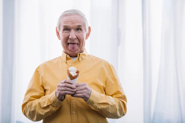 Senior homme qui sort la langue, regardant la caméra et tenant un cône de crème glacée à la maison avec espace de copie — Photo de stock