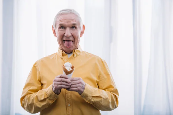 Hombre mayor sacando la lengua, mirando a la cámara y sosteniendo cono de helado en casa con espacio para copiar — Stock Photo