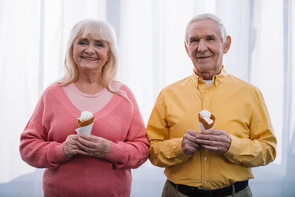 Sonriente pareja de ancianos mirando a la cámara y sosteniendo los conos de helado en casa - foto de stock