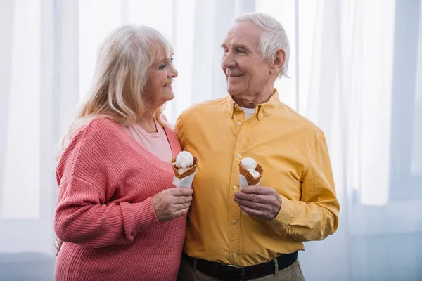 Pareja de ancianos mirándose y sosteniendo conos de helado en casa - foto de stock
