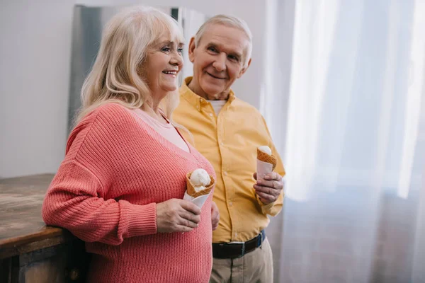 Sonriente pareja de ancianos sosteniendo conos de helado en casa con espacio de copia - foto de stock