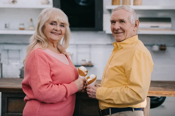 Улыбающаяся пожилая пара смотрит в камеру и держит мороженое конусы дома — стоковое фото