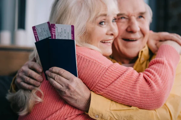 Sonriendo pareja mayor abrazando y sosteniendo boletos aéreos con pasaportes en casa - foto de stock