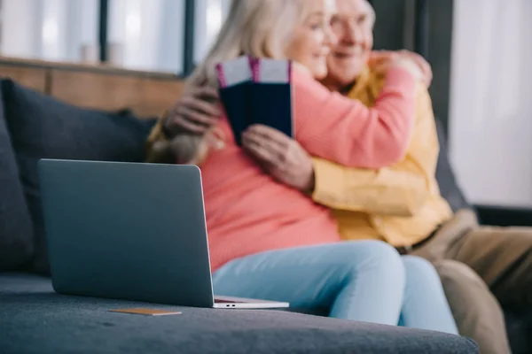 Selektiver Fokus von Laptop und Kreditkarte mit einem älteren Paar, das sich umarmt, während es Flugtickets und Pässe im Hintergrund hält — Stockfoto