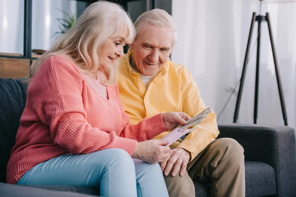 Feliz pareja de ancianos sosteniendo sobre con dinero mientras está sentado en el sofá en casa - foto de stock