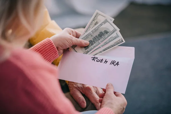 Cropped view of senior couple holding envelope with 'roth ira' lettering and dollar banknotes — Stock Photo