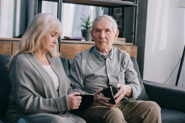 Upset senior couple sitting on couch and holding wallets at home — Stock Photo