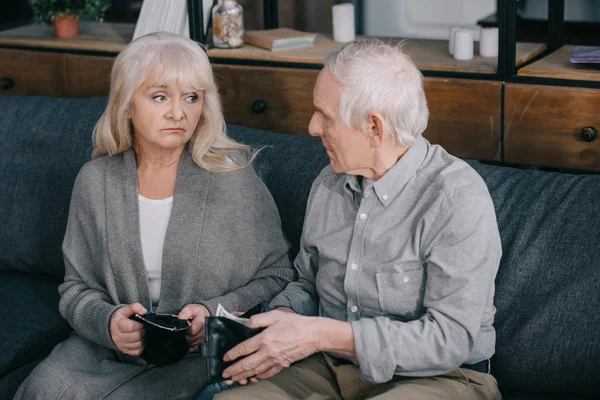 Upset senior couple sitting on couch and holding wallets at home — Stock Photo