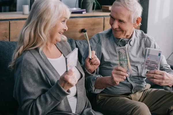 Счастливая пожилая пара сидит на диване и держит банкноты евро и доллара — стоковое фото