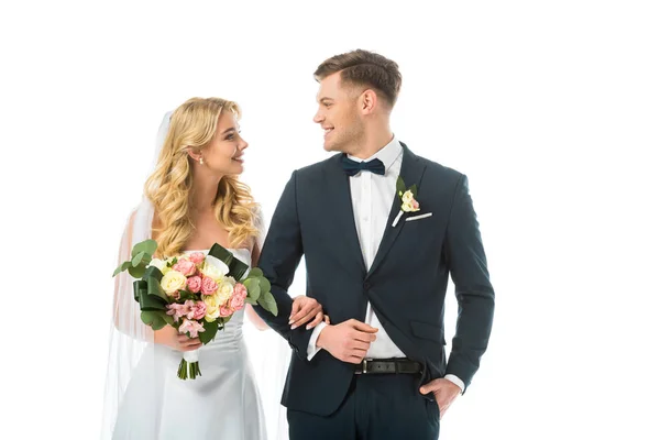 Novia sonriente con ramo de boda, y novio feliz en traje negro mirándose aislados en blanco - foto de stock