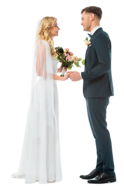 Novia sonriente en vestido de novia blanco y novio feliz en traje negro tomados de la mano y mirándose aislados en blanco - foto de stock