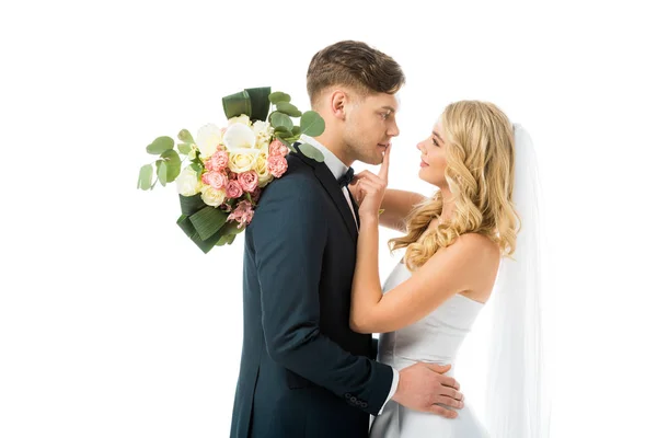 Schöne Braut umarmt glücklichen Bräutigam und zeigt Schweigezeichen auf seinen Lippen isoliert auf weiß — Stockfoto