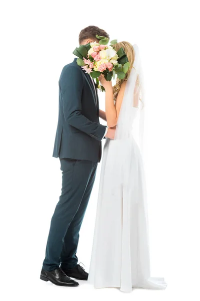 Mariée en robe de mariée longue, et marié en costume élégant embrassant tout en cachant les visages derrière le bouquet de mariage isolé sur blanc — Photo de stock