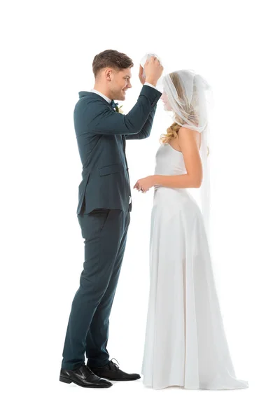 Noivo feliz levantando véu nupcial e olhando para noivas rosto isolado no branco — Fotografia de Stock