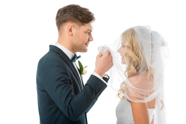 Happy groom looking at smiling brides face while lifting bridal veil isolated on white — Stock Photo