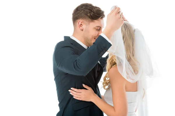 Noivo feliz levantando véu nupcial e olhando para noivas rosto isolado no branco — Fotografia de Stock