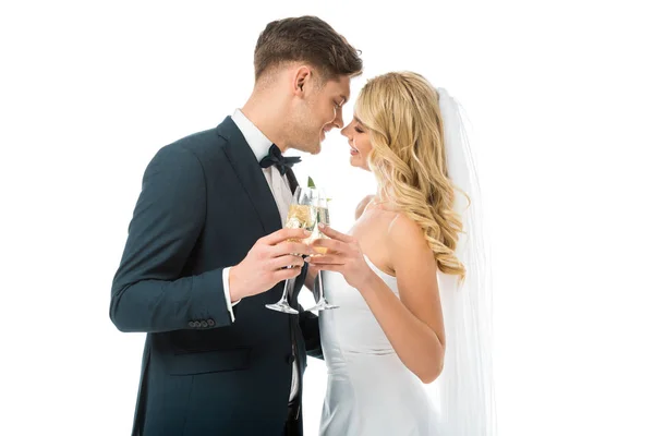 Happy bride and groom clinking glasses of champagne while standing face to face isolated on white — Stock Photo