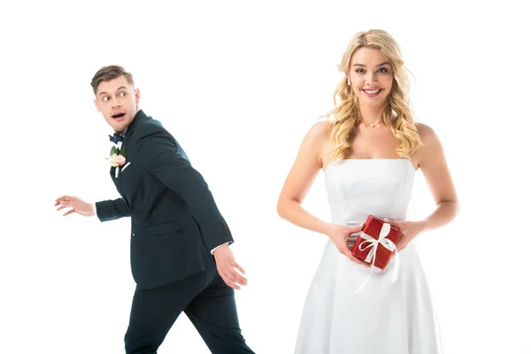 Happy beautiful bride holding gift box, while tricky groom imitating running away isolated on white — Stock Photo