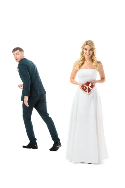 Smiling pretty bride holding gift box, while tricky groom imitating running away isolated on white — Stock Photo