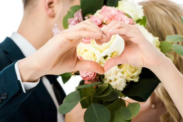 Selective focus of groom and bride showing heart sign with hands, while hiding faces behind wedding bouquet isolated on white — Stock Photo