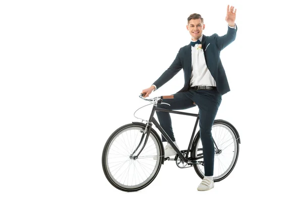 Cheerful groom in elegant suit and sneakers sitting on bike and waving hand isolated on white — Stock Photo