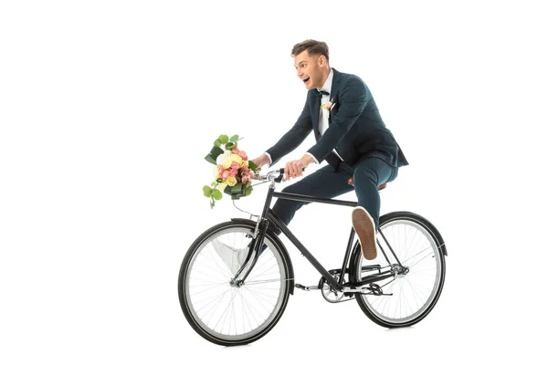 Щасливий наречений в елегантному костюмі і кросівки їзда на велосипеді, тримаючи весільний букет ізольовано на білому — стокове фото