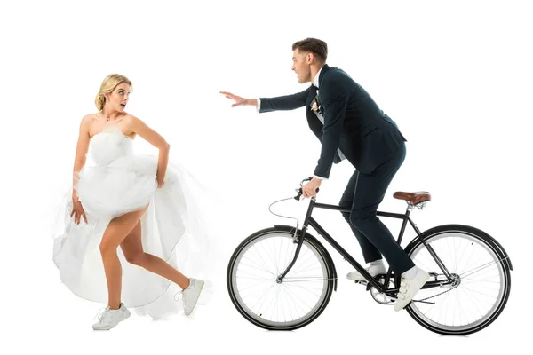 Hübsche Braut im Brautkleid und Turnschuhen, die vom Bräutigam auf dem Fahrrad auf Weiß laufen — Stockfoto
