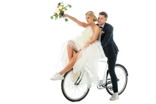 Beautiful bride holding wedding bouquet in raised hand while sitting on bicycle together with groom isolated on white — Stock Photo