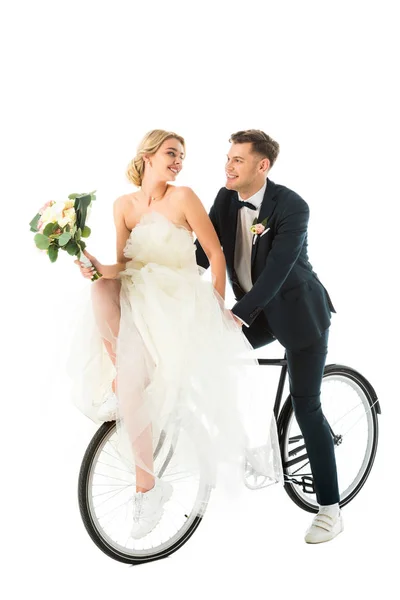 Щаслива наречена з весільним букетом сидить на велосипеді разом зі усміхненим нареченим ізольовано на білому — стокове фото