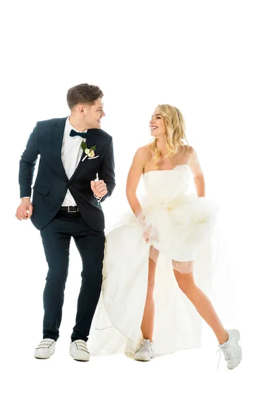 Happy groom and bride dancing in elegant clothes and sneakers while looking at each other isolated on white — Stock Photo