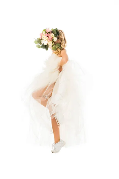 Attraente giovane donna in posa in abito da sposa e scarpe da ginnastica, mentre si nasconde faccia dietro bouquet di nozze isolato su bianco — Foto stock