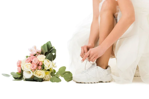 Vista recortada de la mujer atando cordones de zapatos en zapatillas de deporte, y ramo de boda aislado en blanco - foto de stock