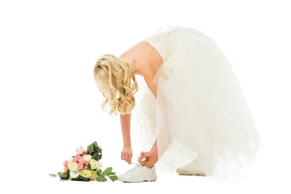 Hermosa novia en vestido de novia atando cordones de zapatos en zapatillas aisladas en blanco - foto de stock