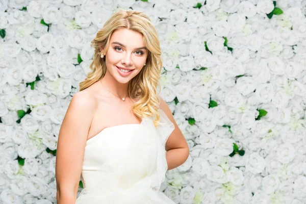 Bella giovane sposa con i capelli biondi guardando la fotocamera su sfondo floreale bianco — Foto stock