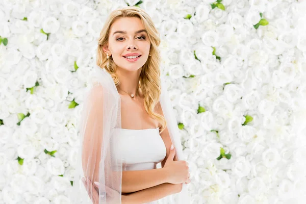 Noiva bonita feliz com cabelo loiro posando na câmera no fundo floral branco — Fotografia de Stock