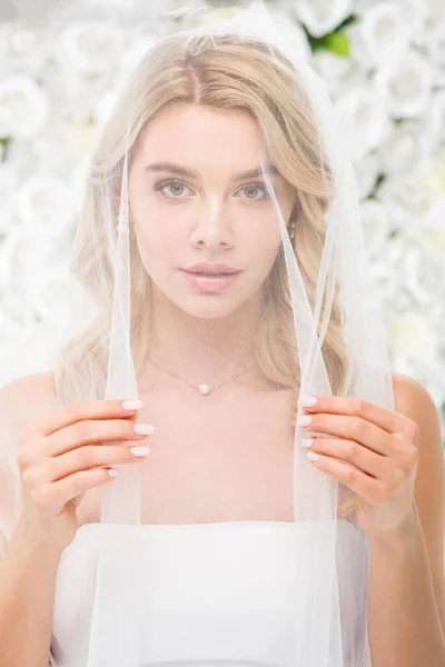 Schöne junge Frau mit Gesicht, das mit transparentem Brautschleier bedeckt ist und in die Kamera auf weißem Blumenhintergrund blickt — Stockfoto
