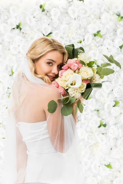 Beautiful young bride holding wedding bouquet near face on white floral background — Stock Photo