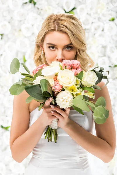 Attractive young bride holding wedding bouquet near face on white floral background — Stock Photo