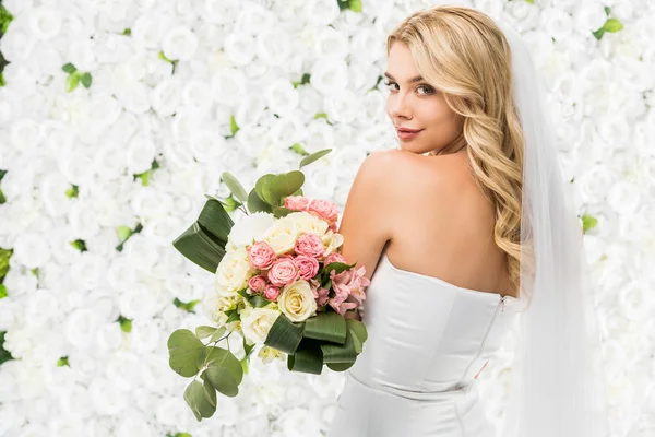 Hermosa novia joven celebración de ramo de boda y mirando a la cámara sobre fondo floral blanco - foto de stock