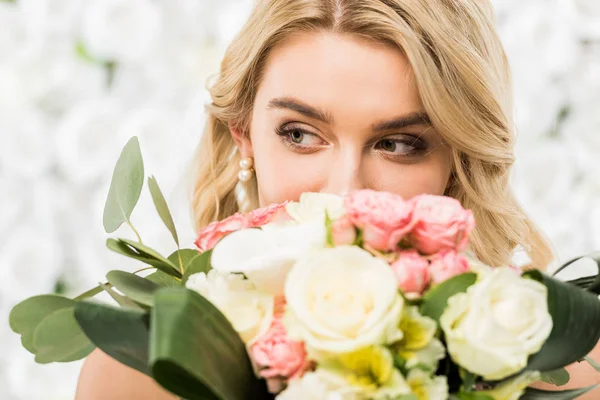 Foyer sélectif de jolie jeune mariée appréciant la saveur du bouquet de mariage sur fond floral blanc — Photo de stock