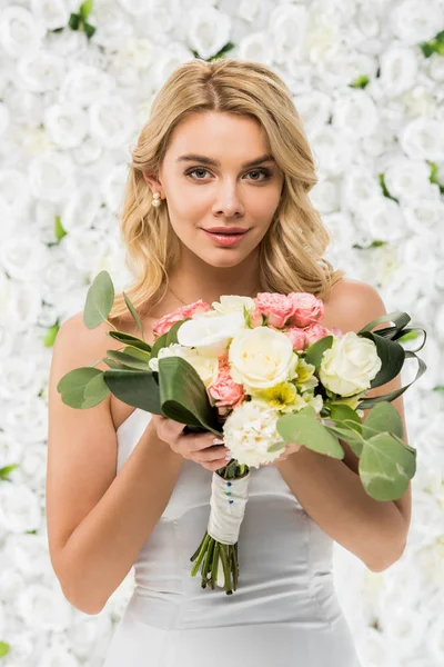 Beautiful young woman holding wedding bouquet and looking at camera on white floral background — Stock Photo