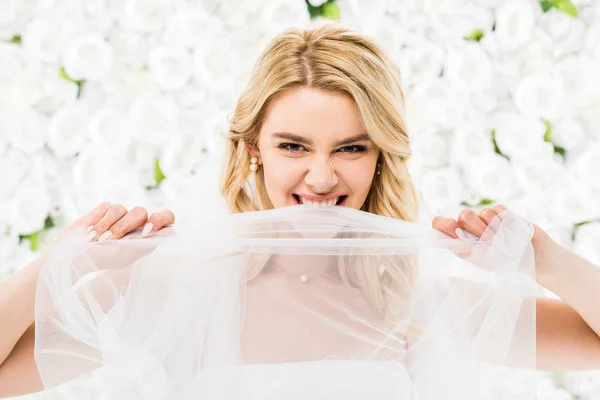 Délicate jeune mariée mordant voile nuptiale sur fond floral blanc — Photo de stock