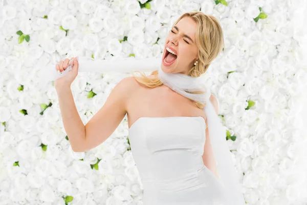 Excited young woman winding bridal veil around neck on white floral background — Stock Photo