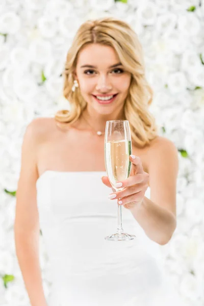 Foco seletivo de sorrir bela noiva segurando copo de champanhe no fundo floral branco — Fotografia de Stock