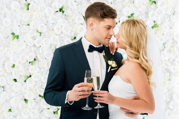 Feliz joven novio y novia de pie cara a cara y sosteniendo copas de champán sobre fondo floral blanco - foto de stock