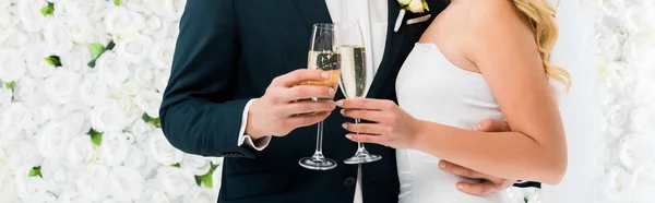 Plano panorámico del novio y la novia sosteniendo copas de champán sobre fondo floral blanco - foto de stock