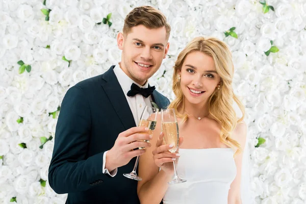 Feliz joven pareja tintineo copas de champán y mirando a la cámara sobre fondo floral blanco - foto de stock